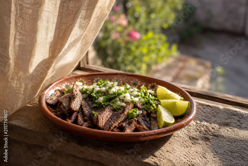 Clay dish of grilled carne asada with cilantro, onion, and lime on stone ledge, authentic Mexican taco filling and food presentation concept