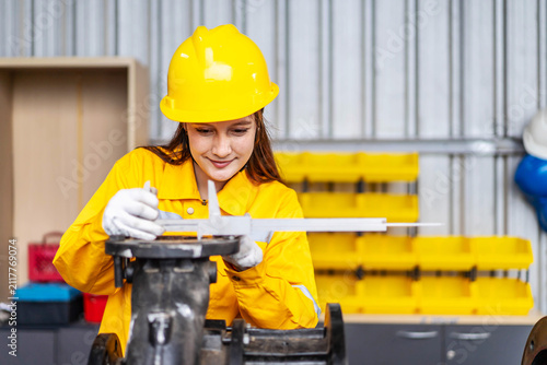 Professional caucasian female engineer wearing yellow safety helmet and uniform using vernier caliper to measure mechanical industrial part component in factory warehouse manufacturing plant workshop