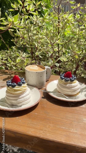 Two pavlova desserts with fresh strawberries and blueberries served with a creamy latte on a sunny outdoor café terrace, creating a cozy atmosphere for breakfast, brunch, or dessert.