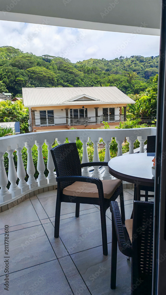 Obraz premium Empty wicker chairs wait on a shaded balcony overlooking a lush tropical hillside, framed from an open doorway with a calm mood. The scene evokes relaxed travel, outdoor living, and peaceful escapes.