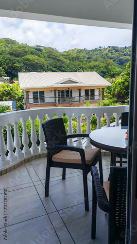 Empty wicker chairs wait on a shaded balcony overlooking a lush tropical hillside, framed from an open doorway with a calm mood. The scene evokes relaxed travel, outdoor living, and peaceful escapes.