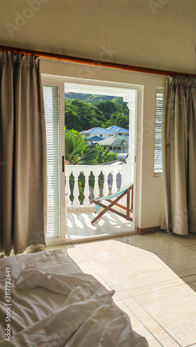 Morning sunlight pours across an unmade bed toward an open balcony, framed vertically from inside a quiet bedroom. The authentic scene evokes slow travel, restful mornings, and tropical living.
