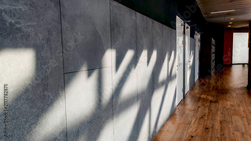 Sunlight casts dramatic geometric shadows along a gray tiled wall in an empty modern corridor, photographed from a low diagonal angle. The scene reflects architecture, minimalism, and urban design.