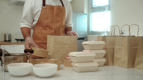 Male chef packaging to paper bags freshly prepared fresh meals for online food delivery in a small business kitchen. Cloud kitchen, local restaurant, food startup. Meal prep and entrepreneur concept.
