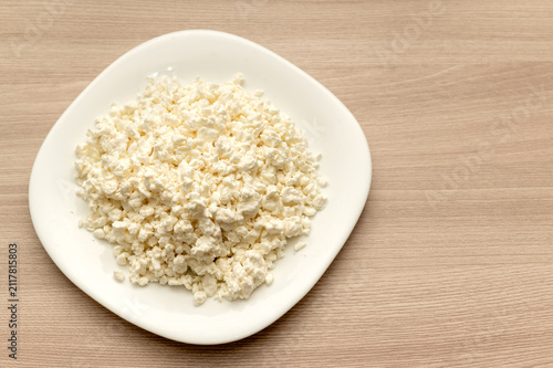 White crumbly cottage cheese on a white plate against a wooden background. Space for text.