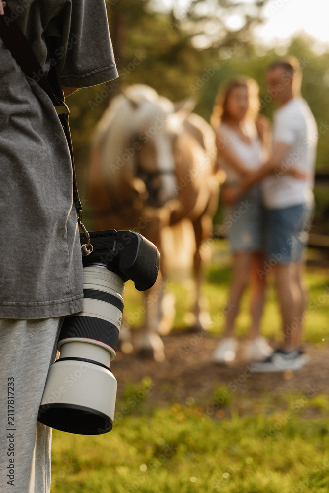 Fototapeta premium Close view of photographer holding large telephoto lens during outdoor horse portrait session, with blurred background horse and person in warm sunlight at relaxed countryside setting
