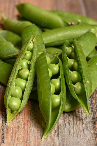 A close-up of fresh green pea pods, some opened to reveal ripe peas, arranged on a rustic wooden surface