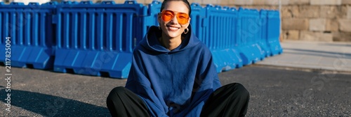 Portrait of woman in oversized blue hoodie with orange mirrored sunglasses sitting cross legged by blue barriers for casual social media banner and background