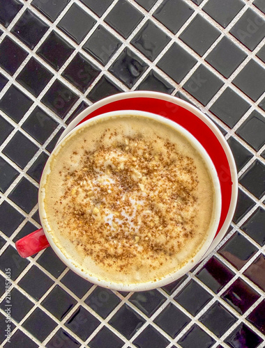 A close-up of a creamy coffee topped with a sprinkle of cinnamon, placed on a black and white tiled surface.