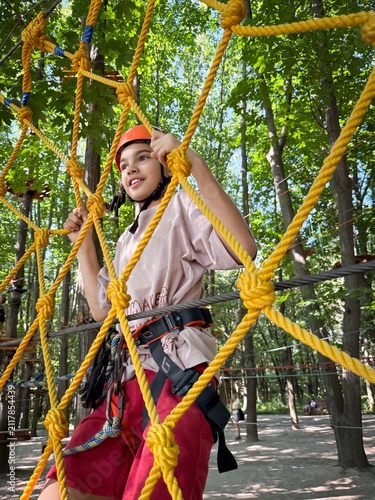 A young girl, of Hispanic descent, navigates an outdoor ropes course, smiling amidst vibrant green trees.