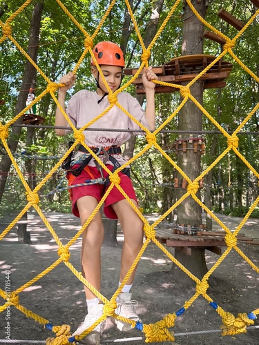 A young girl, possibly Caucasian, in a helmet navigates a climbing net in a vibrant outdoor adventure park.