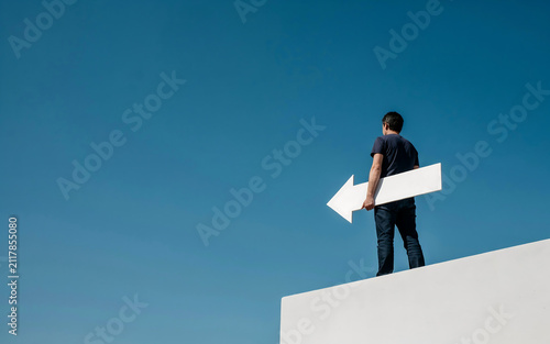 Young man standing on white ledge holding a large white arrow sign pointing left against a clear blue sky with direction decision business navigation pointing strategy opportunity standing