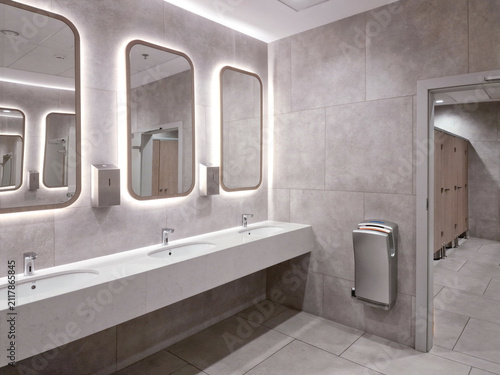 Modern public restroom interior with concrete style tiles, white sinks and illuminated LED mirrors.