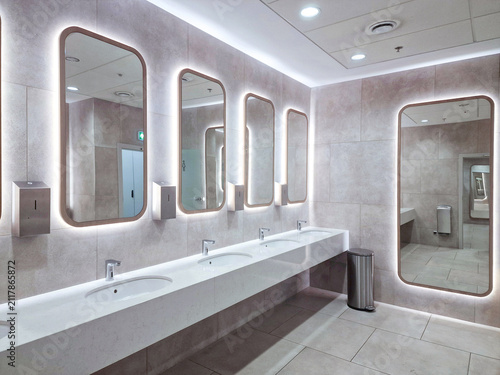 Modern public restroom interior with concrete style tiles, white sinks and illuminated LED mirrors.