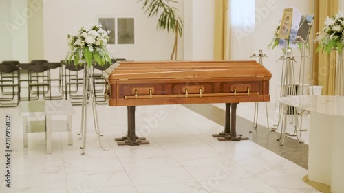 Closed wooden coffin in a bright funeral home or chapel. Solemn farewell ceremony setup with white floral arrangements, empty chairs, and a religious icon.