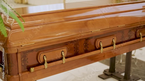 Closed wooden coffin in a bright funeral home or chapel. Solemn farewell ceremony setup with white floral arrangements, empty chairs, and a religious icon.
