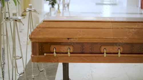 Closed wooden coffin in a bright funeral home or chapel. Solemn farewell ceremony setup with white floral arrangements, empty chairs, and a religious icon.