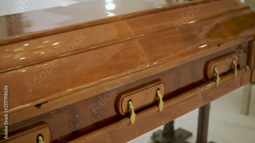 Closed wooden coffin in a bright funeral home or chapel. Solemn farewell ceremony setup with white floral arrangements, empty chairs, and a religious icon.