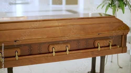 Closed wooden coffin in a bright funeral home or chapel. Solemn farewell ceremony setup with white floral arrangements, empty chairs, and a religious icon.