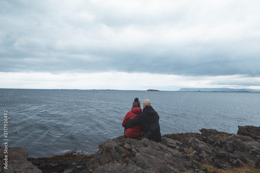 Fototapeta premium Couple sitting together on a rocky Icelandic coast and looking across the cold ocean
