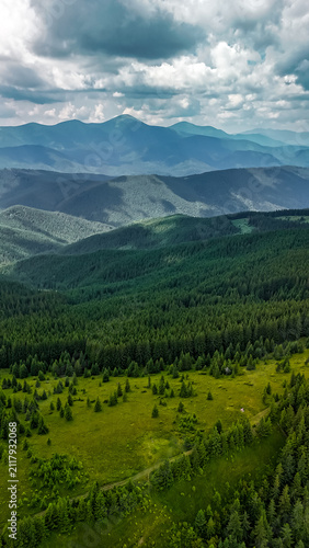 Vertical aerial frame shows layered highlands fading into mist beyond dark spruce slopes. Sunlight slides between clouds lighting meadows and creating a path through wild terrain.