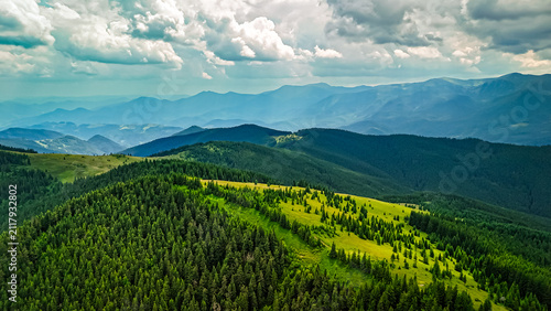 Layers of misty peaks extend to the horizon while forests blanket the foreground. Breaks in the cloud deck reveal meadows and a sinuous ridge that guides the eye across the scene.