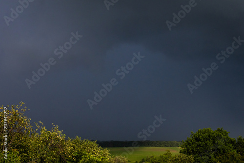 Fotografie Anvil dark clouds dominate the sky above rolling fields and bright hedgerows The