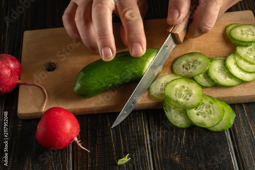 Hands are cutting cucumber on a wooden board while slices are piled next to a red radish. This scene takes place in a home kitchen during meal prep