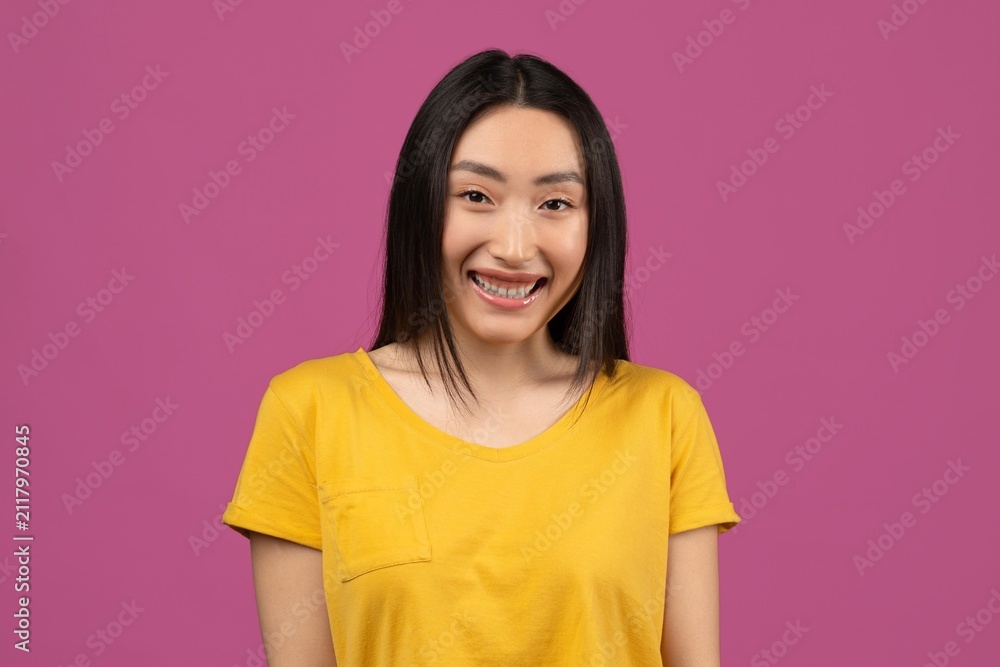 Fototapeta premium Happy Young Asian Woman With Braces Smiling At Camera, Wearing Yellow Shirt, Standing In Front Of Pink Background. Showing Joyful Emotion In A Fun, Colorful Setting. Lifestyle Concept.