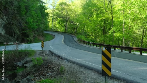 a view of a narrow mountain road, surrounded by tall green trees that create shade in places