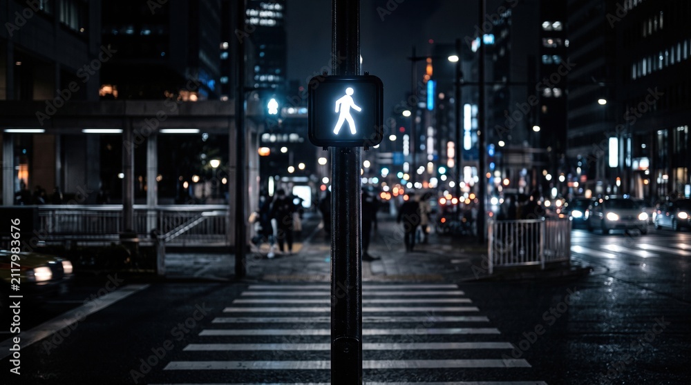 Fototapeta premium White pedestrian walk signal glowing at night on dark city street with blurred people.