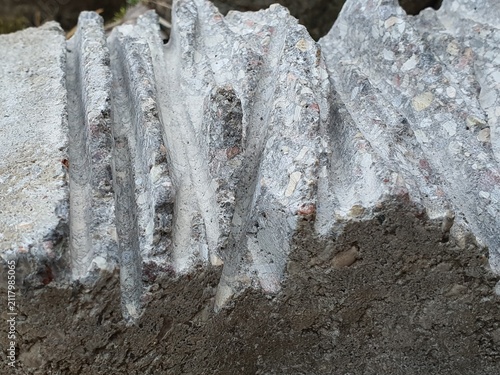 Close-up view of weathered concrete or cement material featuring distinct vertical corrugated ridges and exposed aggregate composition. The rough textured surface displays various embedded stones,