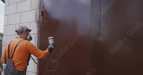 Professional worker in overalls and a respirator using an airbrush to spray paint a metal garage door brown, performing outdoor renovation and surface coating for corrosion protection.