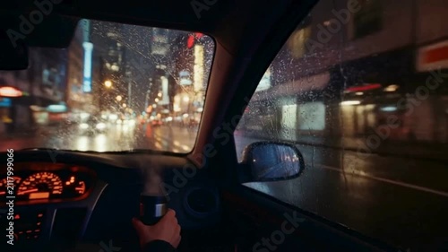 POV driving a car holding a steaming travel mug through a rainy city street at night with neon lights.