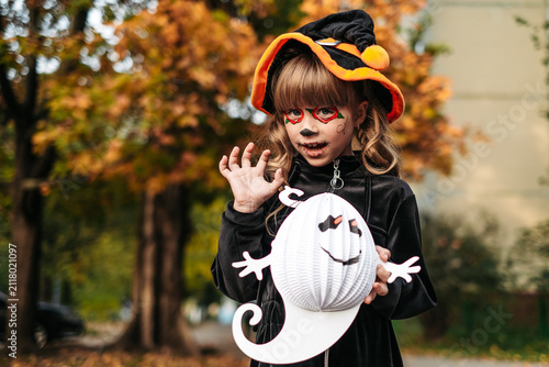 Little girl in witch hat and face paint holding paper ghost outdoors