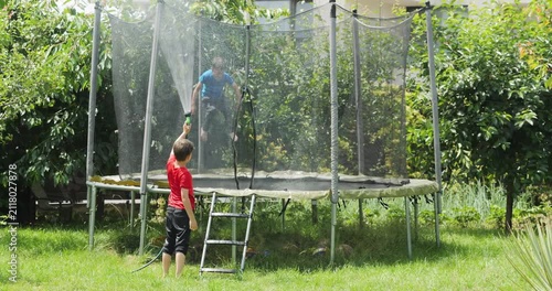 Kid spraying water from garden hose to his brother jumping in trampoline, summer fun