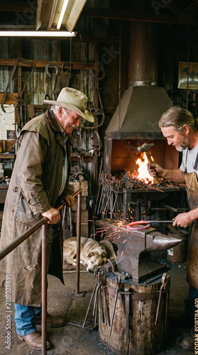 Two men working in rustic blacksmith workshop with anvil, hammer, and glowing hot metal while dog rests nearby, creating warm and focused atmosphere