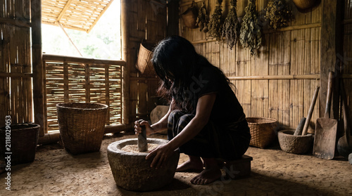 Woman grinding grains in rustic bamboo house with natural light, traditional basket and dried herbs hanging on wall, evoking peaceful rural lifestyle