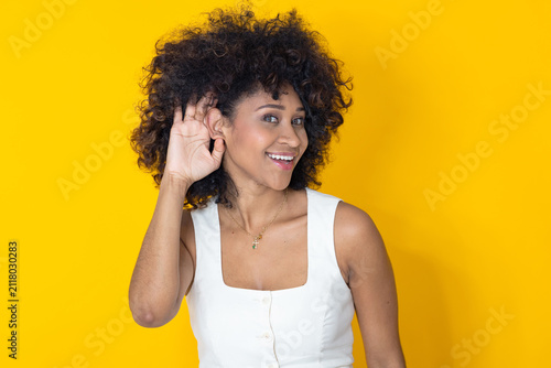 Obraz na plátně Young woman with curly hair cupping her hand behind her ear, smiling and listening carefully against