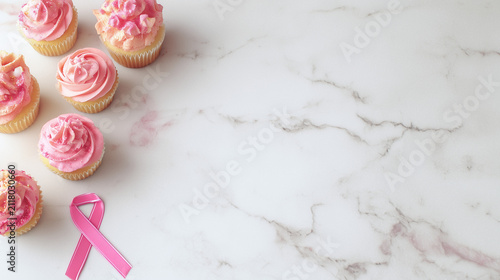 Pink ribbon brooch alongside vanilla cupcakes topped with pink icing on a marble countertop, bakery celebration theme