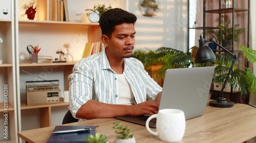 Indian man on home table types on laptop with focused face and steady hands during remote job in living room. Arabian guy on home table works from home with calm mood and productive freelance routine.