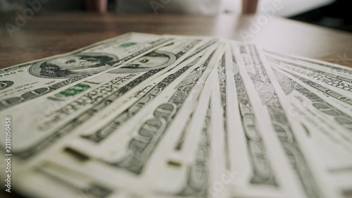 Man slides a fan-shaped arrangement of one hundred dollar bills across a wooden table toward the camera. Visual concept of cash, wealth, financial success, spending, and money management.