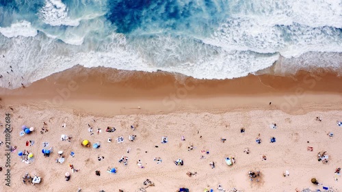 People relax on the beach under umbrellas aerial view. Some swim in the ocean while others walk along the shore. The sun shines brightly on this busy summer day.