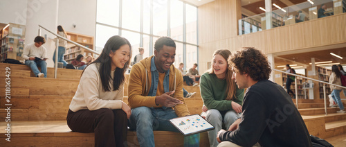 College students studying together in library using a digital tablet with visual data while sitting on wooden campus steps in warm light for university recruitment media