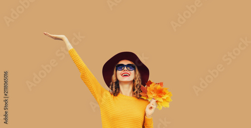 Autumn portrait of happy smiling young woman holding yellow leaves, stylish cheerful girl in sweater