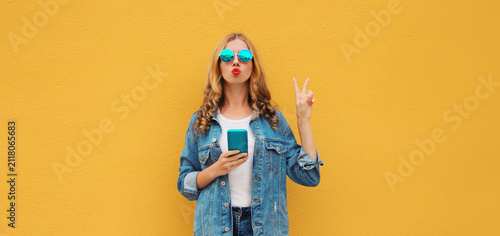 Stylish woman posing blowing a kiss with smartphone in denim jacket casual clothing posing on yellow