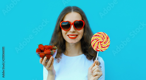 Portrait of beautiful caucasian young woman with handful of fresh strawberries holding sweet lollipop wearing red heart shaped sunglasses on blue studio background