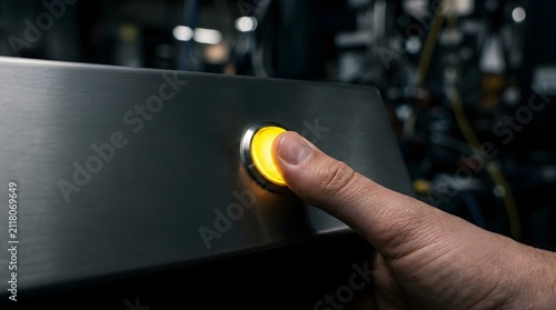 Human finger pressing a glowing yellow industrial power button in a factory