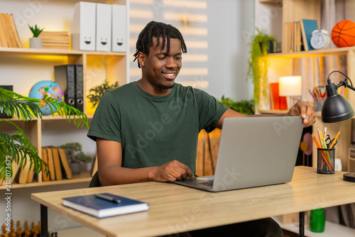 African American young man on home table opens laptop then starts focused remote work with calm smile in living room. Black guy on home table types email using computer for online job freelance task.