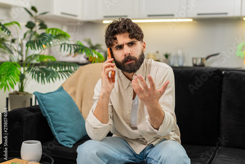 Принти на полотні Indian man at home looks at smartphone gets spam call feels annoyed frowns ends call quickly